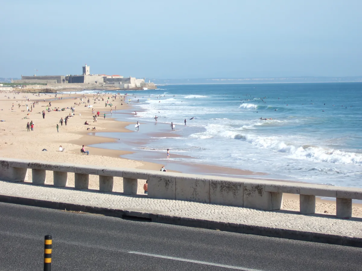 Praia de Carcavelos on a warm day, beachgoers and surfers along the sand below the São Julião fort