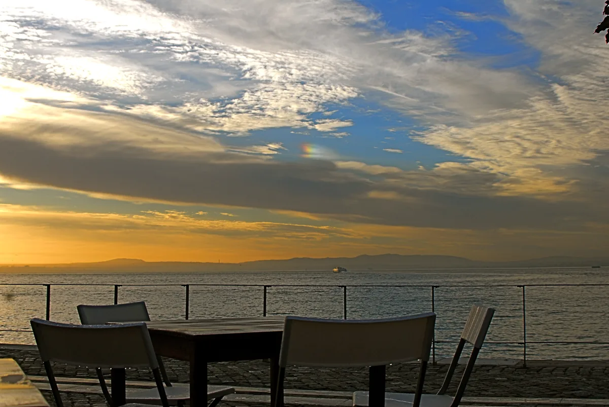 Café chairs on the Tagus riverside under a low winter sunset, a ferry crossing the river behind the railing